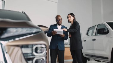 Afrikalı erkek ve kadın, araçlarla çevrili lüks bir galeride, müşteri ilişkileri ve profesyonelliği ön plana çıkaran araba satın alma belgelerini inceliyor.