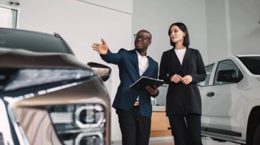 Afrikalı Amerikalı bir adam, çağdaş bir araba galerisinde parlak siyah takım elbiseli bir kadına araba satın alma sürecini ön plana çıkaran bir aracı hevesle sunar.
