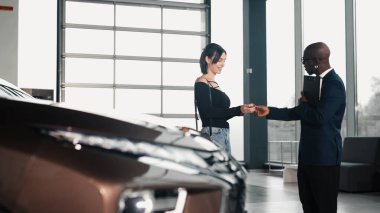 Resmi giyinmiş Afrikalı Amerikalı bir adam, bayide mutlu bir kadının araba anahtarlarını sunuyor. Çağdaş bir ortamda yeni bir otomobil almanın zevkini vurguluyor.