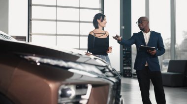 Erkek araba satıcısı kadın müşteriyle yakınlaşır, çağdaş bir bayilik ortamında araç özelliklerini vurgular, araba satın alma yolculuğunu ve müşteri memnuniyetini vurgular