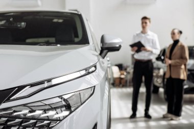 Beyaz SUV çağdaş bir araba galerisinde belirgin bir şekilde sergilenirken, bir erkek satıcı kadın bir müşteriyle temas kuruyor, araba satış sürecini ve etkileşimini vurguluyor