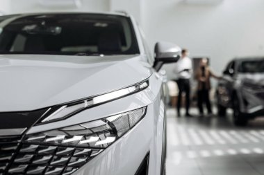 Modern beyaz araç bir galeri galerisinde sergilenirken, lüks tasarımını ve gelişmiş özelliklerini vurgularken, müşteriler satın alma seçenekleri hakkında konuşuyorlar.