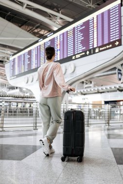 Bavullu kadın gezgin havaalanı terminalinde uçuş bilgisi görüntüleyerek, modern tasarım ve hareketli seyahat ve macera atmosferini vurguluyor