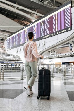 Bavullu bir kadın havaalanı terminalinde uçuş bilgilerini inceliyor, modern tasarım ve geniş bir çevre sunuyor, yaklaşan seyahat macerasına hazırlanıyor.