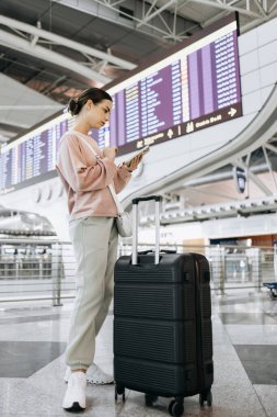 Elinde bavul olan bir bayan gezgin havaalanı terminalinde akıllı telefonunu kullanıyor. Uçuş bilgileri ve arka planda parlak tasarım unsurları var.