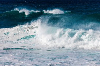 Okyanus dalgaları enerjiyle kumlu kıyıya çarpar. Beyaz köpük ve derin mavi suyun çarpıcı bir görüntüsünü oluşturur. Doğanın gücünü ve güzelliğini gösterir.
