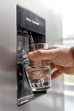 El, temiz bir bardağı temiz suyla doldurmak için bir su haznesinin altına yerleştirilir ve modern mutfak aletlerinin ve su tesisatının kolaylığını vurgular.