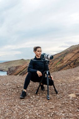 Kadın fotoğrafçı kayalık zemine çömelmiş, nefes kesen manzarayı yakalamak için tripodun üzerinde bir kamera kullanıyor, seyahat ve keşfe olan sevgisini yansıtıyor.