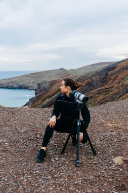 Kadın fotoğrafçı kayalık arazide, çarpıcı kıyı manzaralarını yakalamak için tripodu kullanıyor, doğanın güzelliğini ve keşiflerin neşesini gösteriyor.