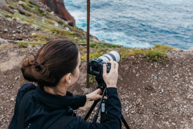 Kadın fotoğrafçı, engebeli kayalar ve okyanus dalgalarıyla çevrili kamerasıyla nefes kesici sahil manzarasını yakalamaya odaklanmış durumda.