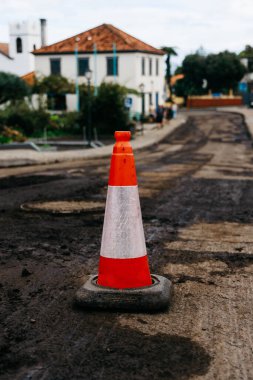Traffic cone stands prominently on a muddy road, with buildings and greenery in the background, signaling ongoing construction and roadwork activities