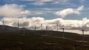 Rüzgar türbinleri bulutların altındaki yeşil yamaçta duruyor.