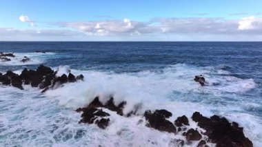 Dalgalar mavi gökyüzünün altında kayalık kıyılara çarpıyor.