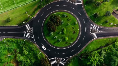 Yeşil peyzaj ve ağaçlarla çevrili bir kavşağın hava perspektifi, huzurlu bir şehir ortamında yol düzeni ve doğal unsurları vurguluyor.