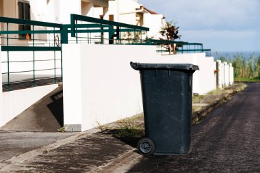 Çağdaş evlerin yanındaki sakin bir sokakta duran çöp tenekesi, kentsel atık imha uygulamalarını ve kamu alanlarındaki temizliğin önemini vurguluyor