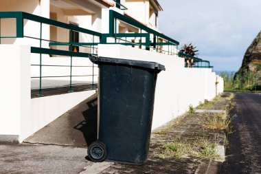 Açık gökyüzünün altındaki bir iskan binasının yanında duran çöp kutusu, kentsel yaşamın ve düzenli bir ortamda atık yönetiminin önemini vurguluyor.