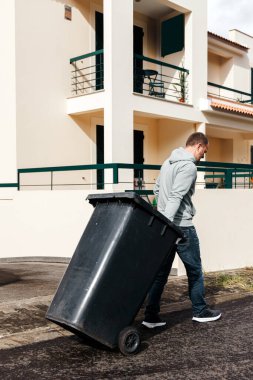 Evinin önünde siyah çöp kutusuyla yürüyen bir adam.