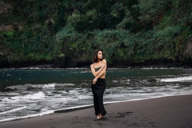 Kadın figürü kumlu sahilde, yalınayak, dalgalar yavaşça kıyıya vuruyor, canlı yeşilliklerle çevrili, huzurlu ve sakin bir ortam yaratıyor.