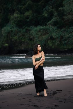 Kadın zarif bir şekilde siyah bir kumsalda duruyor, yeşil bir bluz ve siyah etek giyiyor, arka planda hafif dalgalar var, sakin bir sahil atmosferini yakalıyor.
