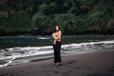 Kadın figürü, dingin, siyah bir kumsalda duruyor. Dalgalar yavaşça yakına çarpıyor. Etrafı canlı yeşilliklerle çevrili.