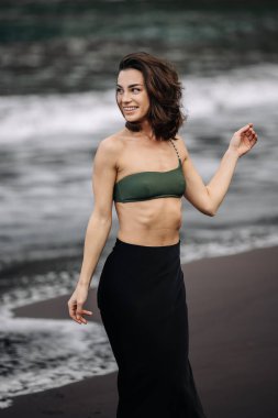 Yeşil bikinili ve siyah etekli gülümseyen genç bir kadın, kumlu sahilde narin okyanus dalgalarıyla yürüyor, kaygısız bir yaz havası ve neşeli bir ruh yakalıyor.