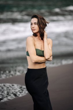 Kendine güvenen genç bir kadın kumlu sahilde duruyor, yeşil bir bluz ve siyah etek giyiyor, dalgalar yavaşça arka plana vuruyor, sükunet ve güzellik anı yakalıyor.