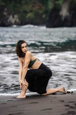 Yeşil bikinili ve siyah etekli bir kadın kumlu sahilde diz çöker, dalgalar yavaşça ayaklarına dokunur, huzurlu bir sahil atmosferi yaratır.
