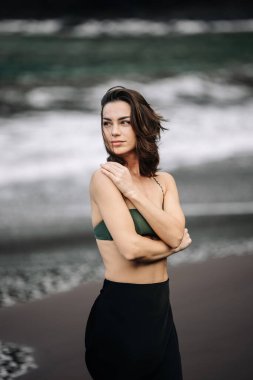 Kadın kumlu sahilde duruyor, okyanus esintisini hissediyor, yeşil bir bikini bluzu ve siyah bir etek giyiyor, doğanın güzelliğinde huzurlu bir anı somutlaştırıyor.