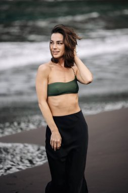 Yeşil bikinili ve siyah etekli kadın figürü kumlu sahilde duruyor, okyanus esintisini hissediyor, dalgalar geliyor, rahatlatıcı bir yaz atmosferi yaratıyor.