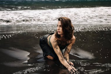 Siyah kumlu plajda diz çöküp vücudunu boyayan ve dalgalarla nazikçe etrafını saran sanatsal genç kadın yaratıcılığı ve doğayla olan bağlantısını somutlaştırıyor.