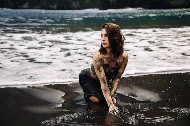 Dişi figür kumlu sahilde diz çöküyor, derisi çamurlu, dalgalar ona çarpıyor, canlı yeşilliklerle çevrili, bir keşif anını ve doğal güzelliği yakalıyor.