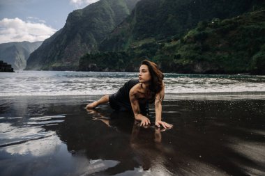 Siyah elbiseli kadın figürü kıyı şeridine zarif bir şekilde yerleştirilir. Dalgalar yavaşça kuma ve dağlara vurur ve dingin bir zemin oluşturur.