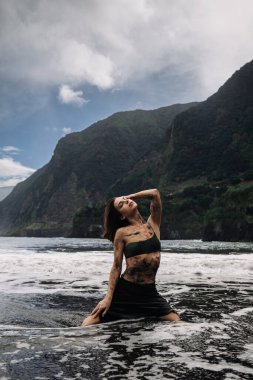 Kadın kıyıda zarifçe poz veriyor, etrafını dalgalar sarıyor, arka planda kayalıklar yükseliyor, sükunet ve doğal güzelliği yakalıyor.