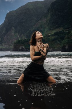 Koyu çamurla süslenmiş, okyanus dalgalarında diz çökmüş, çarpıcı uçurumlar ve yeşillikler arasında, doğa ve kişisel yolculukla derin bir bağlantıyı simgeleyen kadın figürü.