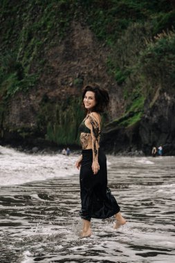 Siyah mayo ve etek giymiş neşeli genç bir kadın, okyanus dalgalarında sıçrıyor, arka planda kayalık kayalıklar ve yeşillikler, canlı bir yaz atmosferini yakalıyor.