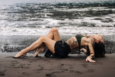 Siyah bikinili kadın figürü kumlu sahilde, çamurla süslenmiş, okyanus dalgaları plajda nazikçe tur atarken, sakin ve sanatsal bir atmosfer yaratıyor.