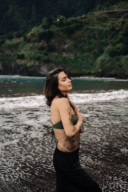 Yeşil bikini ve siyah etek giymiş, kumlu sahilde duran, okyanus esintisini hisseden ve sakin bir kıyı yaşamını kucaklayan bir kadın.