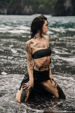 Siyah bikinili kadın figürü kumlu sahilde diz çökmüş, çamurla kaplı, etrafı okyanus dalgalarıyla çevrili, doğa ve sanatsal ifadenin eşsiz bir karışımını sergiliyor.