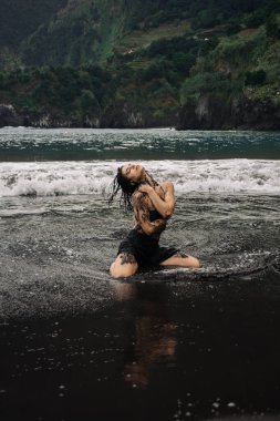 Siyah mayo giymiş kadın dansçı, okyanus dalgalarında neşeli bir şekilde hareket ediyor. Etrafına su sıçrıyor, canlı yeşil tepelere vuruyor, neşe ve özgürlüğü ifade ediyor.