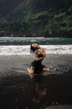 Dövmeli bir kadın, okyanus dalgalarında diz çökmüş, doğayla bağını ve özgürlüğünü somutlaştırıyor, dramatik uçurumlar ve sakin sahil manzaralarıyla çevrili, bir güç hissi uyandırıyor.