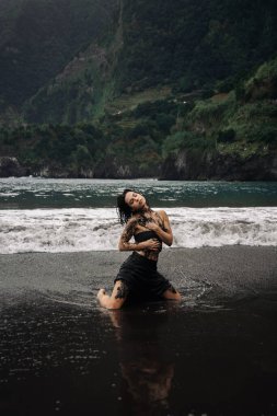 Dövmeli kadın figürü, siyah kumsalda poz veriyor, etrafını çevreleyen dalgalar, canlı yeşil tepeler, doğanın özünü ve kendini ifade edişini yakalıyor.