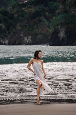 Zarif beyaz elbiseli kadın sahilde duruyor, dalgalar ayaklarını okşuyor, arkada kayalıklar ve okyanus var, huzurlu ve huzurlu bir atmosfer yaratıyor.
