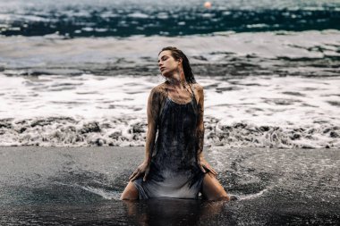 Kum üzerinde diz çökmüş, dalgalarla çevrili bir kadın figürü, doğal bir sahil ortamında huzur ve yoğunluğun bir karışımını somutlaştırıyor.