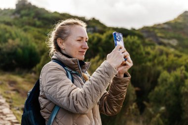Dişi yürüyüşçü, çevresi tepeler ve ağaçlarla çevrili yemyeşil bir arazide fotoğraf çekmek için akıllı telefon kullanıyor. Açık hava keşiflerinin neşesini sergiliyor.