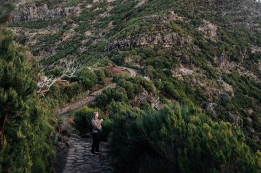 Dişi gezgin, canlı yeşillikler ve kayalık tepelerle çevrili dolambaçlı bir yolda duruyor. Huzurlu bir atmosferde büyüleyici manzaralar yakalıyor.