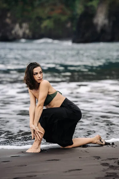 Yeşil bikinili ve siyah etekli bir kadın kumlu sahilde diz çöker, dalgalar yavaşça ayaklarına dokunur, huzurlu bir sahil atmosferi yaratır.