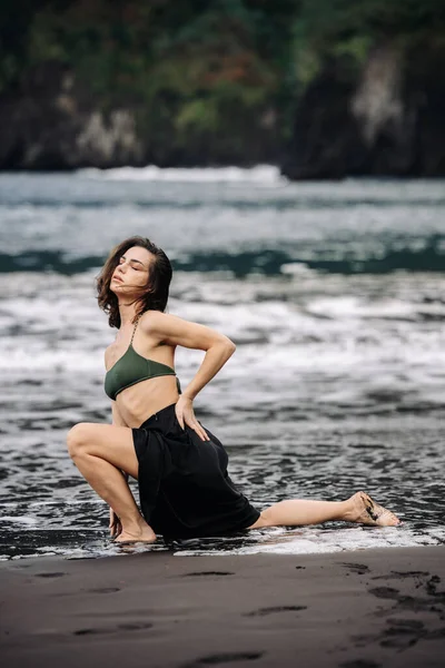 Yeşil bikinili ve siyah etekli bir kadın, okyanus kıyısındaki ıslak kumda diz çöker, doğayla uyum ve sükuneti simgeler, pitoresk bir plaj ortamında.