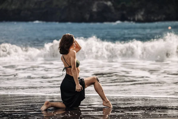 Kumlu sahilde diz çökmüş bir kadın, bikini ve etek giyiyor, okyanus dalgaları yakınlarda çarpışırken, güneş ışığı ve doğal güzelliklerle dolu huzurlu bir atmosfer yaratıyor.