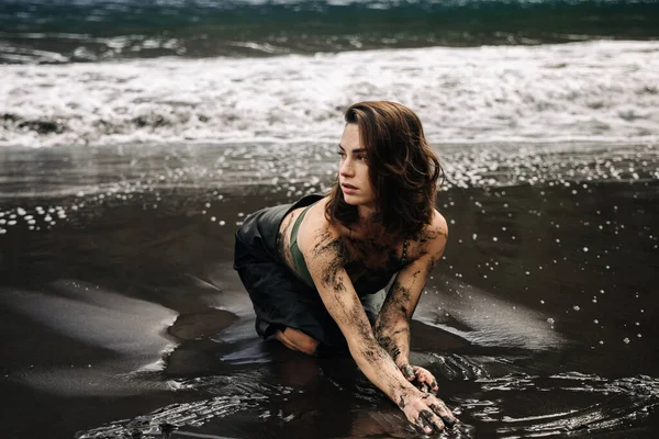 Siyah kumlu plajda diz çöküp vücudunu boyayan ve dalgalarla nazikçe etrafını saran sanatsal genç kadın yaratıcılığı ve doğayla olan bağlantısını somutlaştırıyor.