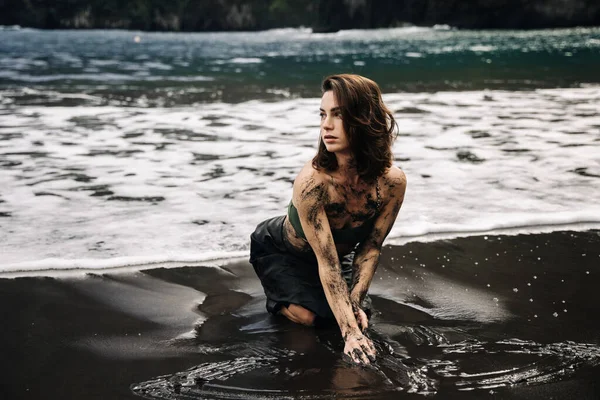 Dişi figür kumlu sahilde diz çöküyor, derisi çamurlu, dalgalar ona çarpıyor, canlı yeşilliklerle çevrili, bir keşif anını ve doğal güzelliği yakalıyor.